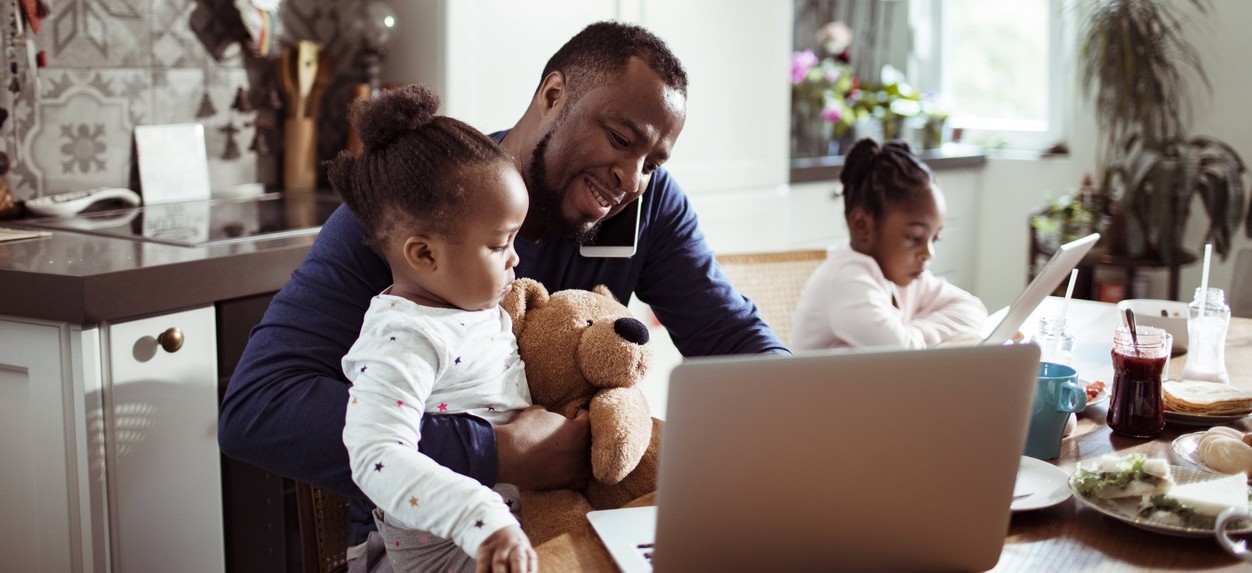Father at table on computer & phone with one child on lap and other next to him.