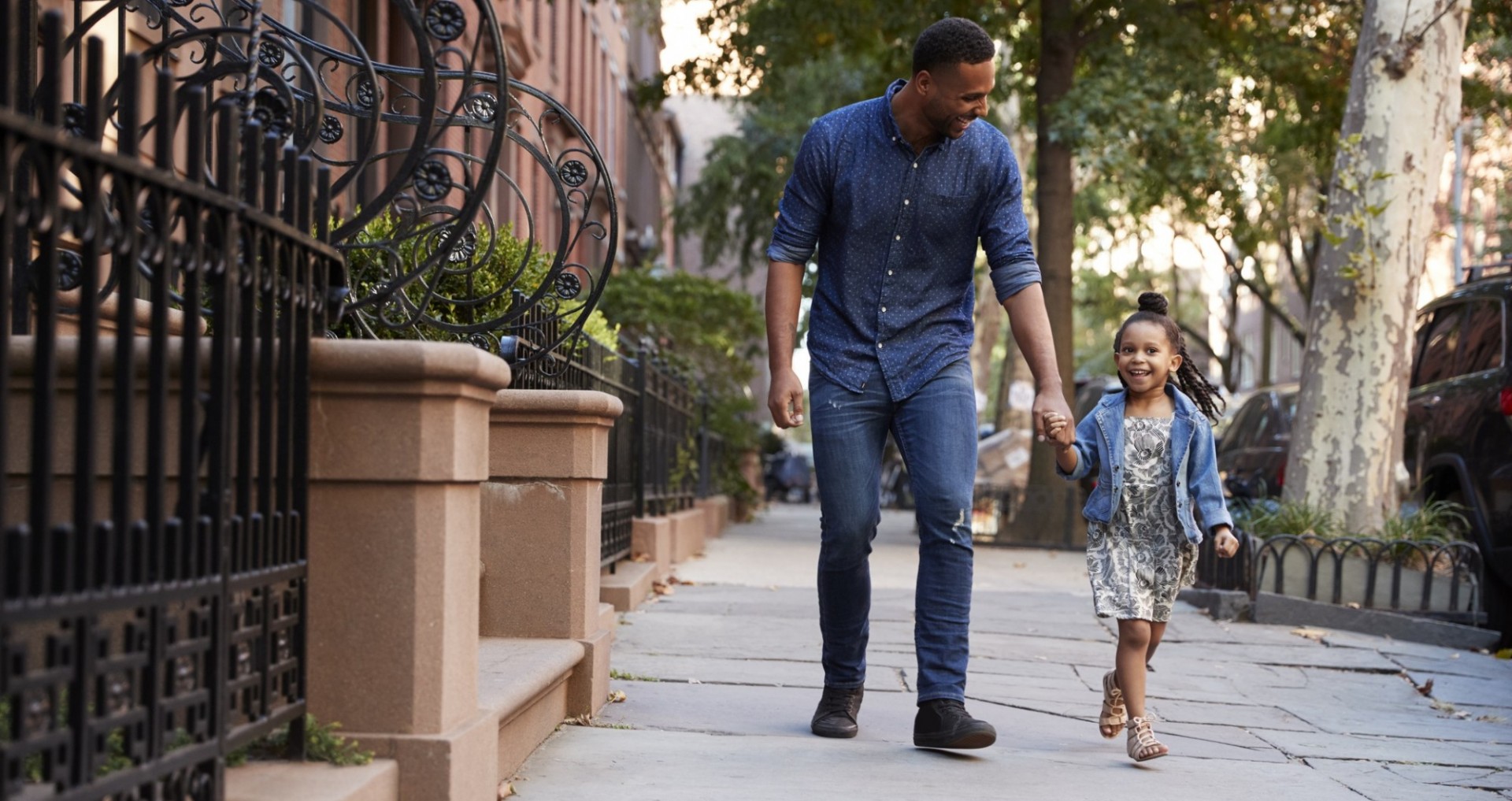 Father daughter holding hands walking down city tree-lined street.