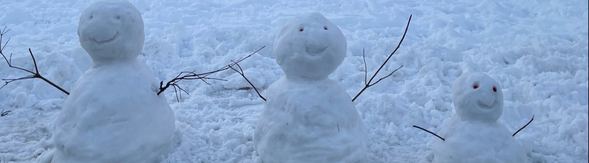 Three smiling snow people with stick arms on snow-covered ground.