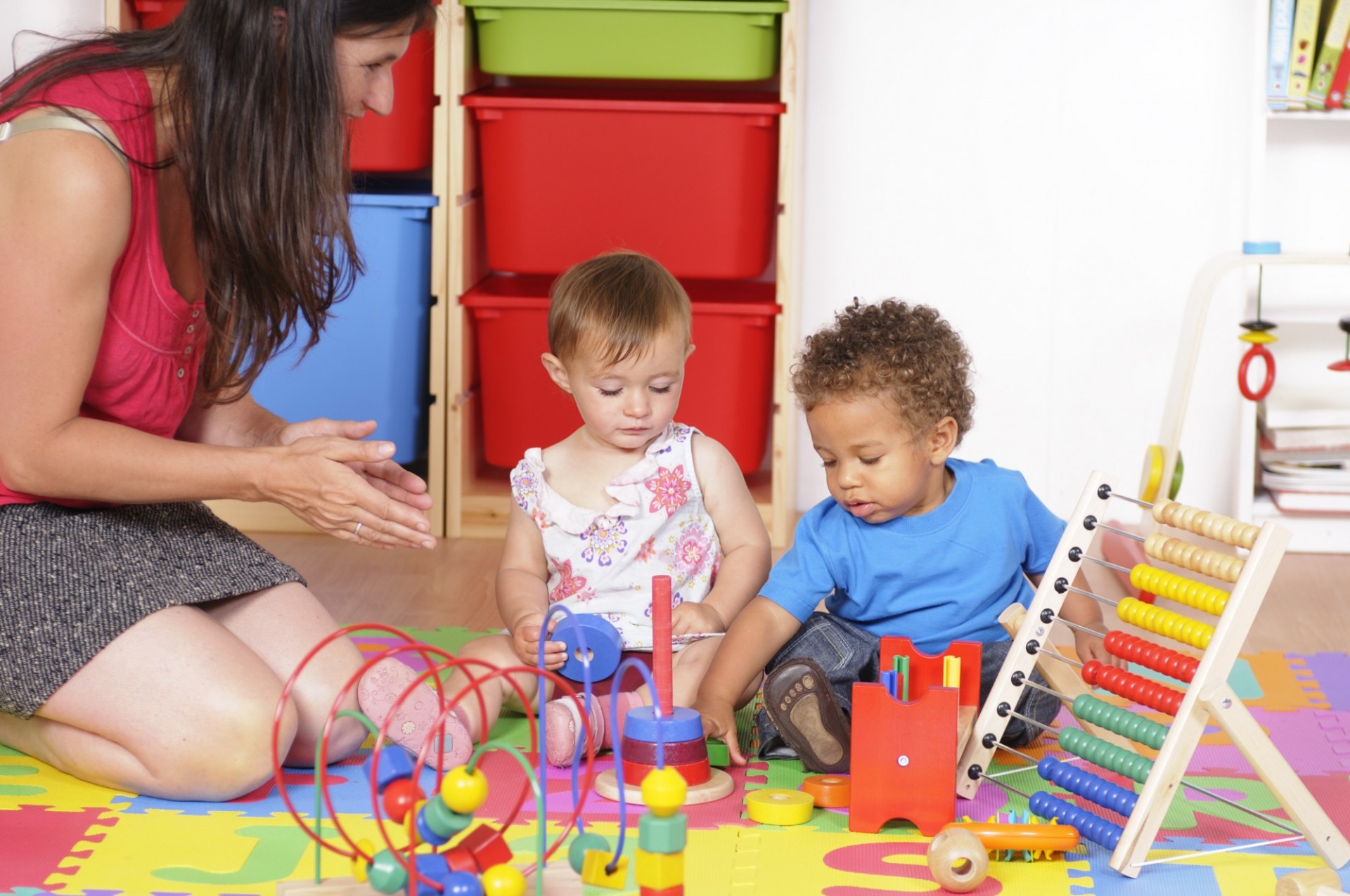 Two toddlers playing on floor with brightly colored toys while adult looks on.