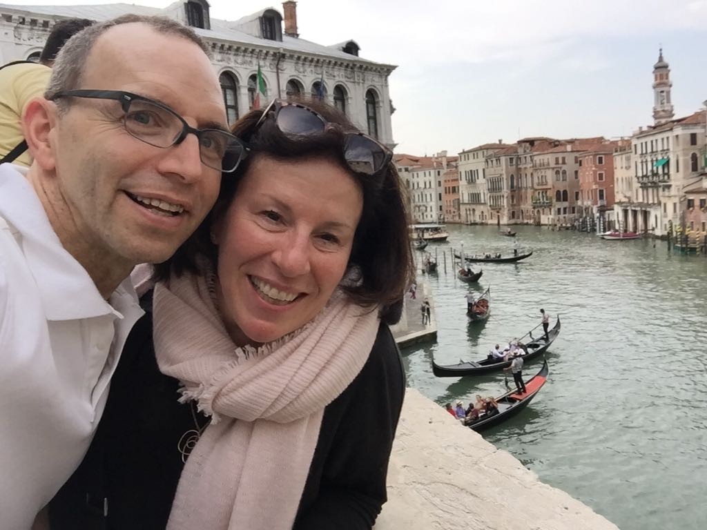 smiling man and woman with venice buildings, canals, and gondals in background.