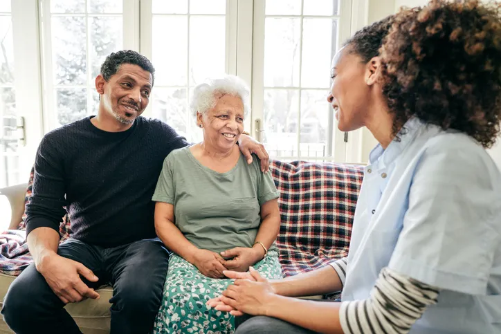 Older white-haired woman sitting side-by-side with younger man on couch, talking with woman sitting adjacent.