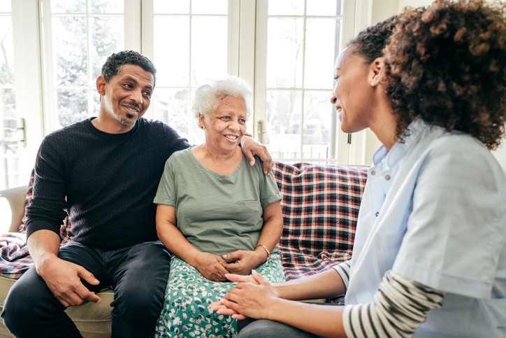 Older white-haired woman sitting side-by-side with younger man on couch, talking with woman sitting adjacent.