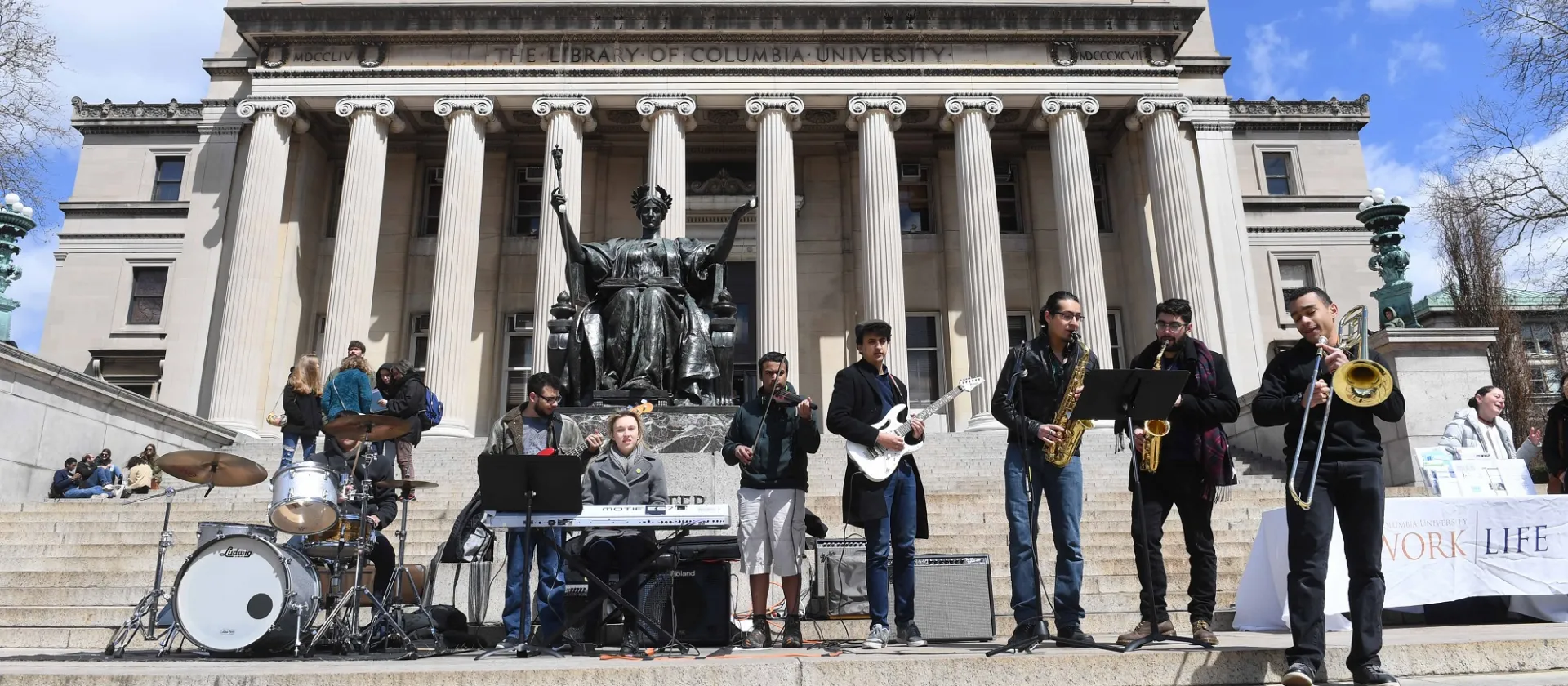 Bank playing various instruments on large steps in front of columned building with blue sky overhead.
